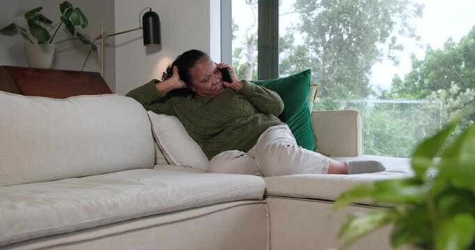 Senior African American woman taking call while lying on couch with phone, laughing, catching up