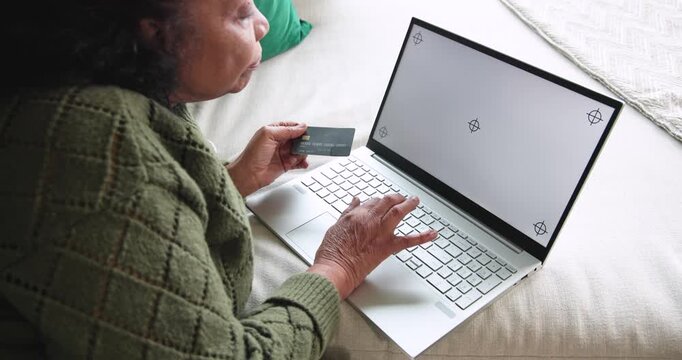 Senior African American woman in green knit holding card, typing on laptop on sofa entering payment