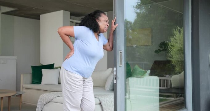 Senior African American woman holding lower back seeking relief, leaning on door frame at home