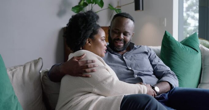 African couple sitting on couch at home, woman touching man's wristwatch, showing love, smiling