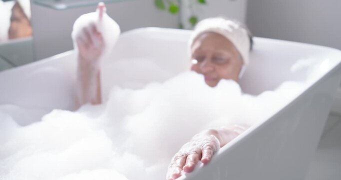 Senior African American woman listening to music to relax via earbuds, scooping foam in bathtub