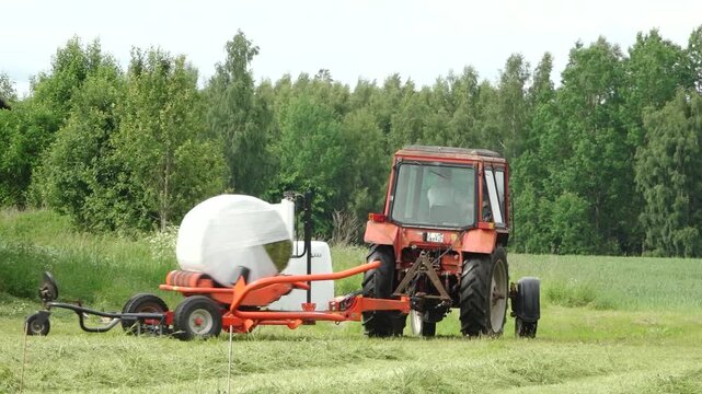 Old tractor pulls a bale wrapper across a cut grass field, wrapping a round hay bale in white plastic with dense green forest in the background. Plani, Latvia - June, 12, 2026.