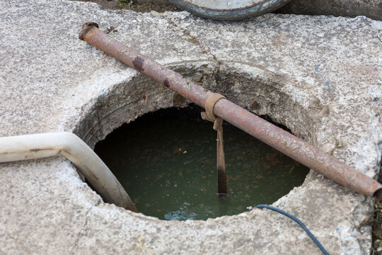 An open concrete well or septic tank is filled with dirty water during drainage or maintenance work. A rusty metal pipe with a tied strap spans across the opening, and a flexible hose is lowered insid