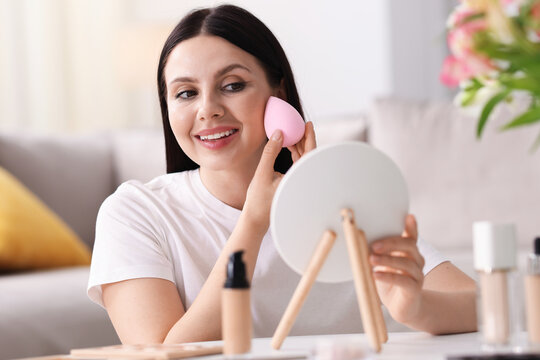 Young woman with mirror applying foundation with makeup sponge at table indoors