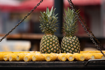 Obraz premium Eye-level shot of two whole pineapples sitting on iron grill rack with corn cobs below, metal chains frame both sides. Blurred red market awning behind creates vivid urban street food atmosphere