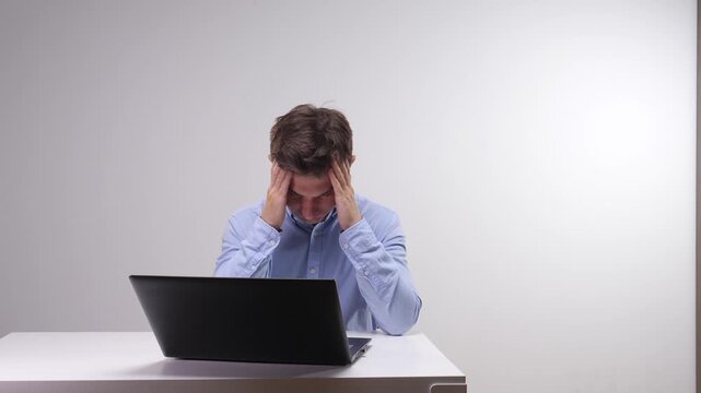 Stressed man sitting at a desk with a laptop, holding his temples, indicating pain and exhaustion from work overload and job burnout