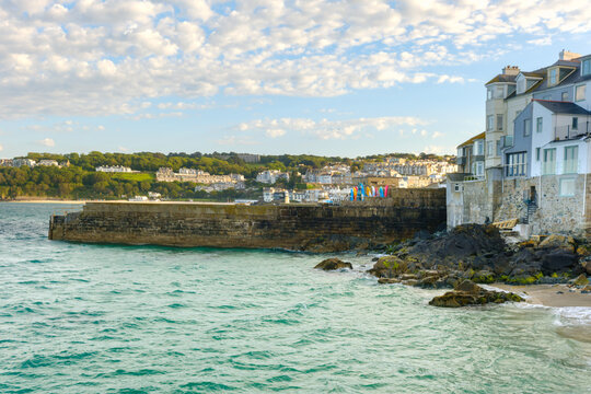 View of the historic stone harbor wall and coastal buildings overlooking the turquoise sea under a cloudy sky in St Ives, Cornwall, United Kingdom.