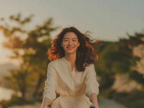 Delighted young East Asian lady in pale yellow flowy blouse cycles outdoors at golden hour, sunlight gilds her fluttering long hair amid softly blurred warm-toned green trees in background.