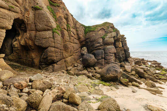 View of the rugged granite cliffs, cave entrance, and natural rock arch at Porthgwarra Beach under a cloudy sky Porthgwarra, Cornwall, United Kingdom.