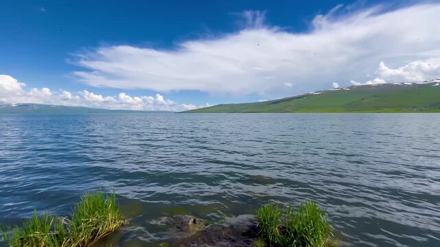 Low-angle POV shot slowly approaching the rocky shore and clear rippling waters of Lake &Ccedil;ıldır (&Ccedil;ıldır G&ouml;l&uuml;) in spring, surrounded by green hills