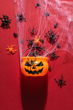 Orange Halloween pumpkin bucket surrounded by plastic spiders and white spiderweb on dark red background. Vertical template for spooky holiday celebration, trick or treat party, or invitation.
