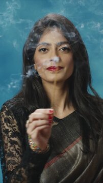 Close up portrait of beautiful woman smelling incense stick and looking forward. Female enjoying aroma and smoke while standing indoors. Traditional outfit and calm ritual moment at home.