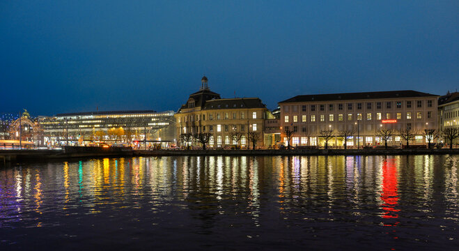 Beleuchtete Geb&auml;ude am Bahnhofsplatz und Seebr&uuml;cke in Luzern bei Blauer Stunde mit Spiegelung