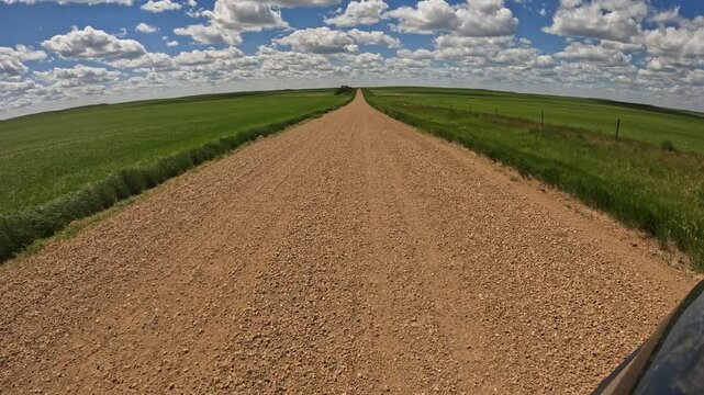 Point of view driving along a rural gravel road through open prairie under a summer sky. Crossing a cattle gate and approaching a park
