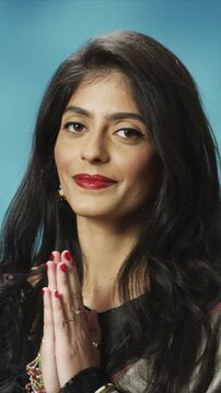 Close up portrait pf beautiful young Indian woman holding hands together in greeting gesture and looking at camera. Female wearing traditional clothing. Cultural expression indoors on blue background.