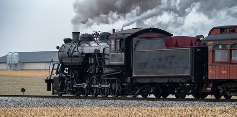 A steam train moves along the tracks in the countryside. Smoke billows from the locomotive as it passes by fields and buildings. The scene shows a typical day of train travel. © Greg Kelton