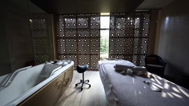 Interior spa room shows massage table and hydrotherapy tub side by side, star pattern screen, dappled light moves on towels, orchids, tassel, warm wood tone, central symmetry.
