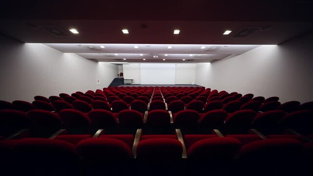 Include title in description. Empty lecture hall with deep red seats in six columns, white projection screen, small stage with side stairs at left, ceiling projector, cool lights, no movement.