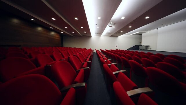 Empty lecture hall with red seats, wood wall, small stage, stairs, projector, and strip lighting. Wide angle view, lights on, slight flicker, projector lens visible.