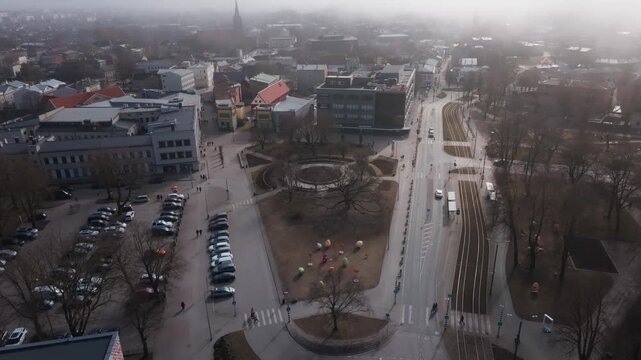 Aerial town center with circular sunken garden and oak, tram tracks, main road, egg sculptures, parked cars, crosswalks, buses, church spire. Include title in description.