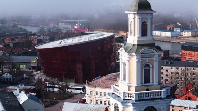Aerial over Liepaja, Latvia shows Lielais Dzintars, baroque church tower, harbor cranes, moored tugboats and vessels, busy streets, cool morning light, gentle mist.