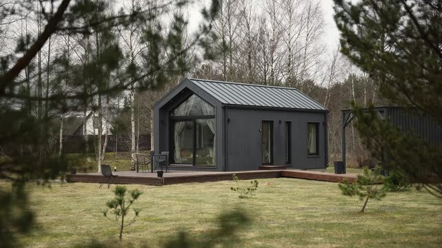 Modern cabin with dark cladding, metal gable roof, and glass frontage sits on a wooden deck among pines and birch. Camera pulls back slowly on an overcast day.