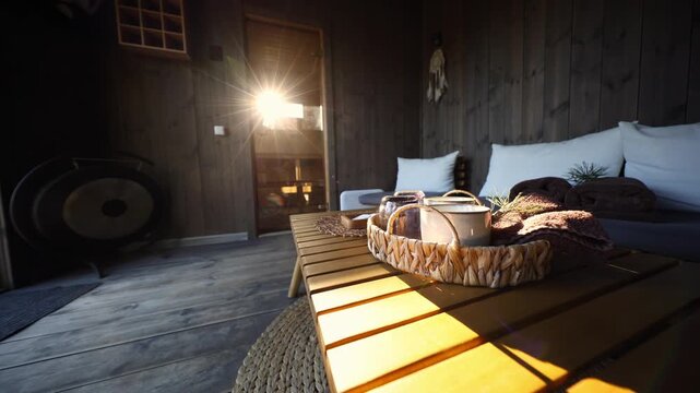 Sunlit spa cabin interior, wood paneled walls, gong on left, dreamcatcher near door, woven tray with cups and towels, bands of light move across bench and rug.