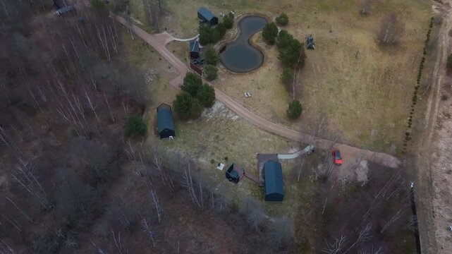 Aerial retreat by birch woodland with figure eight pond and cabins. A slow pan shows dark roofs, an orange car, winding drive, and muted late winter tones.