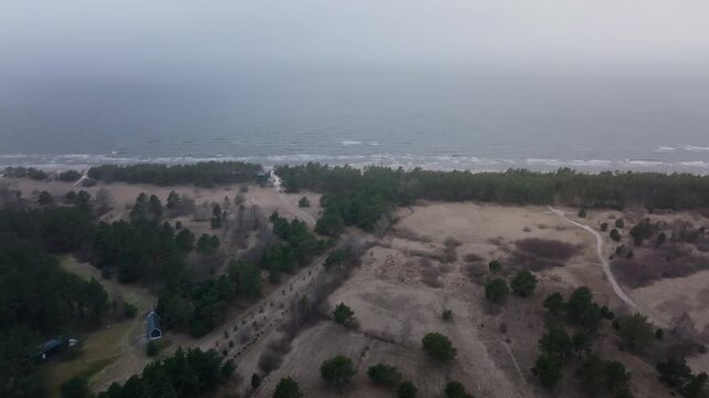 Aerial view shows coastal dune, pine strip, and grey sea as the camera drifts seaward. Rows of wooden groynes, a small cabin, and a dirt track sit under overcast light.