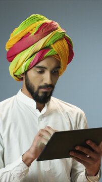 Focused portrait of handsome man wearing colorful turban and holding tablet while reading screen. Male using digital device and concentrating. At home. Indoor modern lifestyle technology scene.