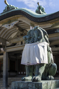 View of a traditional komainu lion-dog statue wearing a white bib in front of a Japanese shrine with a curved roof in Osaka, Osaka, Japan.