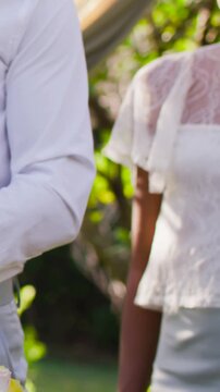 Vertical video: Tilting camera showing couple in white attire readying to cut cake in yard, glass