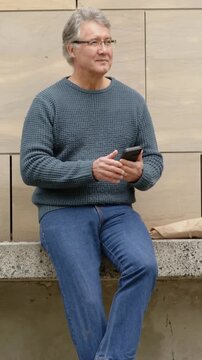 Vertical video: Tapping man in teal sweater checking phone on bench, then looking up with paper bag