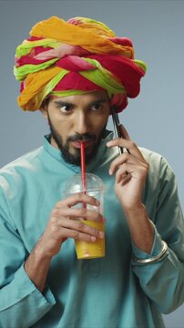 Focused guy, portrait handsome man wearing colorful turban holding smartphone and drinking juice with straw. Male sipping drink and listening to call. Communication. Indoor phone calling moment.