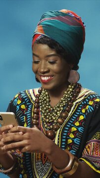 Portrait of African American woman wearing traditional outfit and head wrap using smartphone. Female smiling while texting and reading messages at home. Indoor modern cultural lifestyle scene.