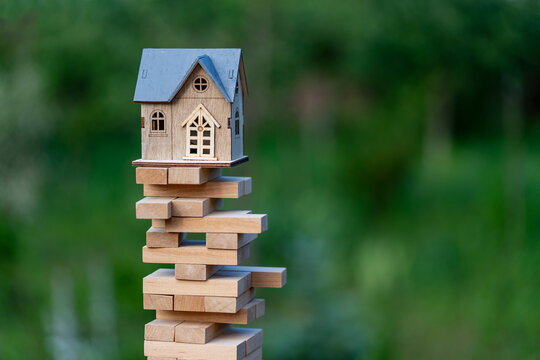 Wooden house model balancing on unstable toy block tower. Concept of real estate risk and financial instability showing a small wooden house model on top of shaky tower with green nature background