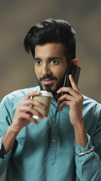 Lifestyle close up portrait handsome man holding smartphone and takeaway coffee cup while listening conversation. Male smiling softly and enjoying break. At home. Indoor daily routine scene.