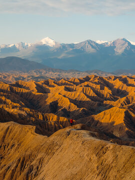 View of a person in a red jacket standing on a ridge in Ak Say Canyon with eroded orange hills and snow-capped mountains in Ak Say, Issyk-Kul Region, Kyrgyzstan.