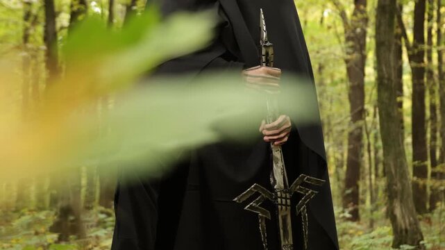 Cloaked figure, woodland setting, forest mood. Close-up cloaked figure in black holding intricately designed sword, set against forest background with blurred leaves.
