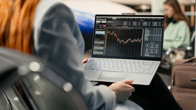 Professional woman reviewing stock charts and financial market insights in airport before departure, using online tools to track algorithms and hedge fund performance, digital nomad freedom.
