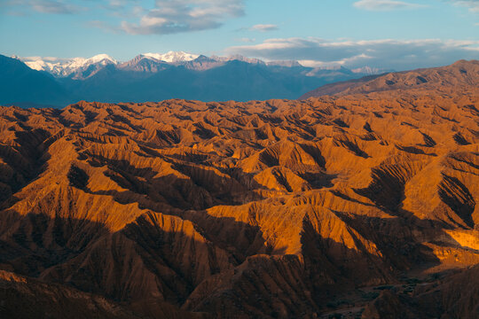 View of the arid eroded hills of Ak Say Canyon with snow-capped mountains in the distance under a soft blue sky, Ak Say, Issyk-Kul Region, Kyrgyzstan.