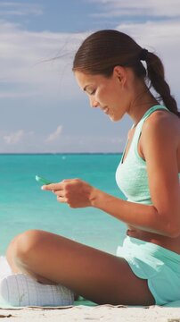 Fit girl using fitness app on phone during travel holidays on beach texting or posting on social media online. Healthy sporty Asian woman living a happy active life exercising on towel. VERTICAL VIDEO