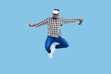 Obraz premium Excited Young Black Man Engaging With Virtual Reality Headset While Jumping In Indoor Studio Setting, Holding Game Controller. Enjoying New Gaming Experience In Modern Environment.