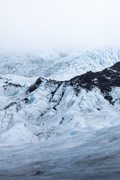 View of the rugged blue ice formations and black volcanic ash of a glacier under a misty sky in Iceland.