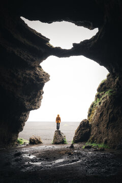 View of a person in a yellow jacket standing on a rock inside a dark volcanic cave looking out at a bright landscape Iceland.