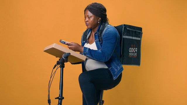 Black woman delivers takeout orders on bicycle, utilizing wireless payment device for seamless transactions. African american delivery person taking pos machine from pocket for contactless pay.