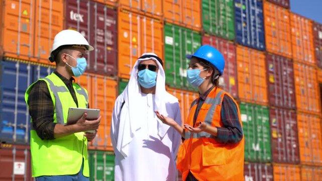 Arab businessman in kandura and logistics workers in masks discussing cargo details with digital tablet in front of colorful shipping containers stock video.