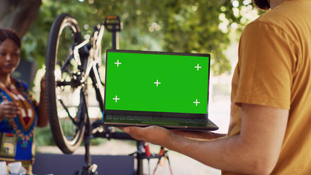 Close-up shot of laptop with isolated chromakey display carried by male cyclist while Black woman dismantles damaged bicycle wheel, with couple doing bike maintenance with minicomputer having green.