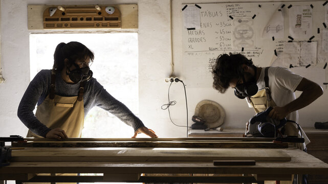 View of a man and a woman wearing protective respirators and aprons while working with a circular saw on a wooden board in a workshop in Lisbon, Lisbon, Portugal.