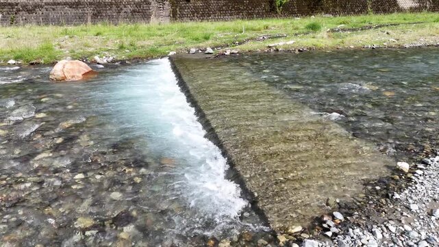 Clear water rushes over a stone barrier. Rocks bed the streambed beneath the current. Single orange stone rests near the flow. Green grass lines the bank behind the water